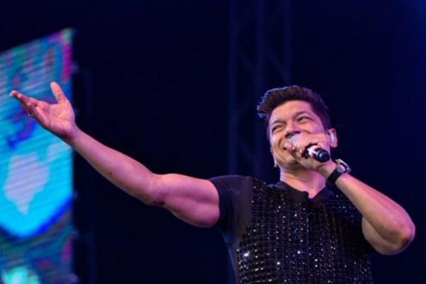 Shaan is singing on stage into a microphone, wearing a sequined black t-shirt.